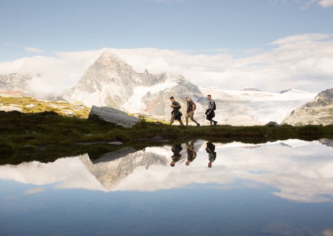 Three hikers walking next to a lake with snowcapped mountains behind them