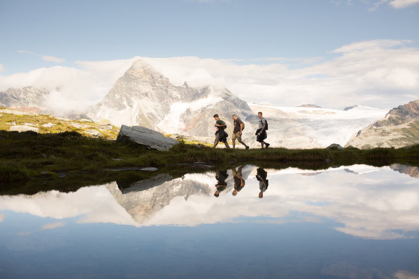 Three hikers walking next to a lake with snowcapped mountains behind them